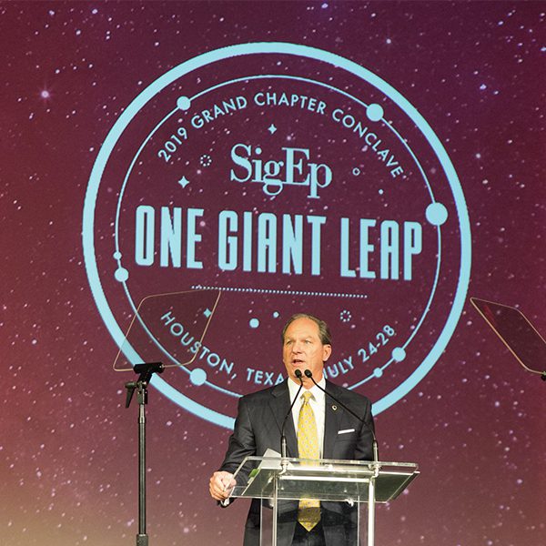 man speaking in front of sigma phi epsilon logo