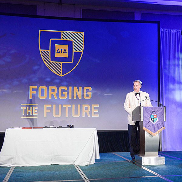 DTDF's new Forging the Future logo is shown projected on a screen while a man stands at a podium giving a speech