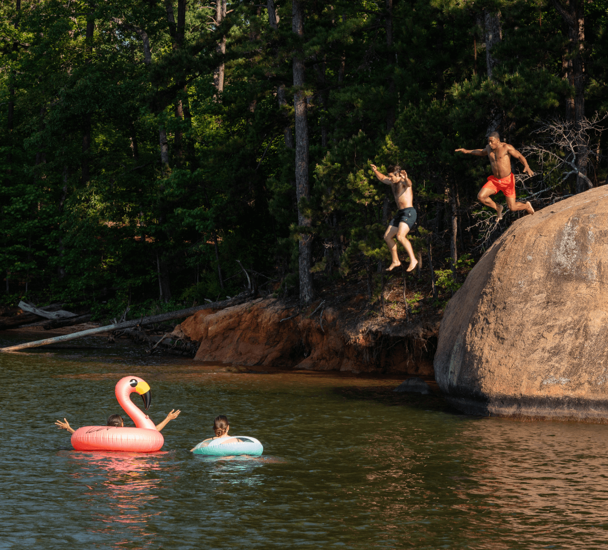 Photo of two friends jumping off a rock into a lake. Below, two more friends are in the water on floaties.